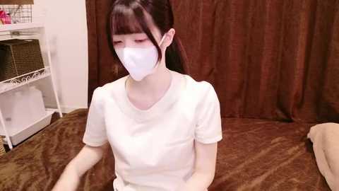 Media: Video of a young Asian woman with fair skin, dark hair, wearing a white mask, short-sleeved white shirt, sitting on a brown bedspread. Background includes white shelves with a wicker basket, and brown curtains.