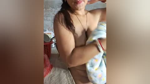 Media: Video of a topless, light-skinned woman with medium-length brown hair, smiling, holding a blue and yellow towel, in a room with a white brick wall and a red plastic laundry basket.