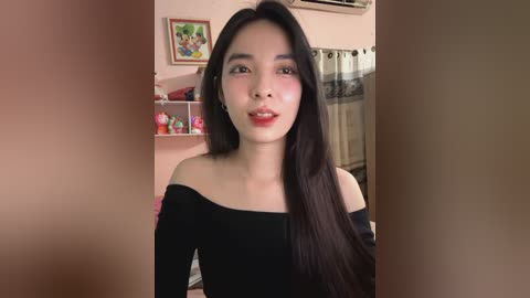 Media: Video of an East Asian woman with long black hair, wearing a black off-shoulder top, smiling in a cozy room with a pink wall, framed art, and a window with a beige curtain.