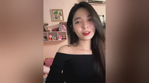 Media: A video of an East Asian woman with long black hair, wearing a black off-shoulder dress, smiling in a bedroom with a pink wall, a shelf filled with plush toys, and a framed picture.