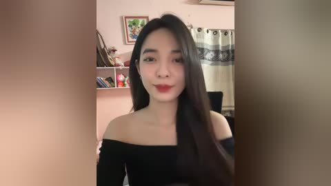 Media: Video of a young Asian woman with long black hair, wearing a black off-the-shoulder top, standing in a cozy room with bookshelves and a curtain.