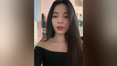 Media: Video of an East Asian woman with long, straight black hair, fair skin, and full red lips, wearing a black off-the-shoulder top, standing indoors with a blurred background of shelves and clothes.