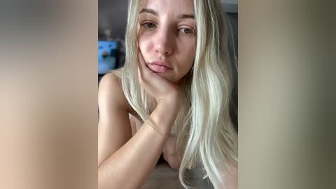 Media: A video of a topless young woman with long, straight platinum blonde hair, fair skin, and light makeup, leaning forward with her chin on her hand, gazing directly at the camera.