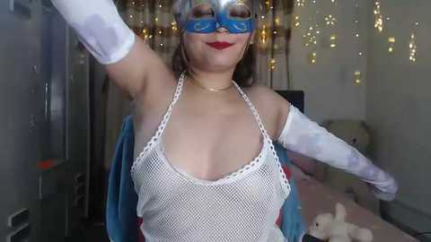 Media: Video of a woman dressed as Wonder Woman in a white fishnet top, blue mask, and cape, standing in a room with fairy lights and plush toys.