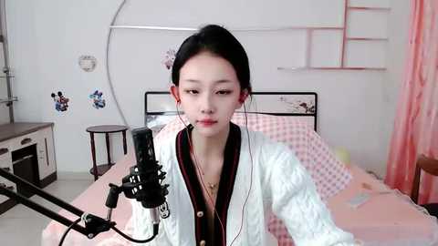 Media: Video of an East Asian woman with pale skin, black hair in a bun, wearing a white cardigan, singing into a microphone in a softly lit, minimalistic bedroom with pink curtains.