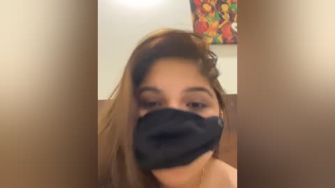 Media: Video of a young woman with light brown hair, wearing a black face mask, capturing her partially obscured face and a colorful, abstract painting in the background.