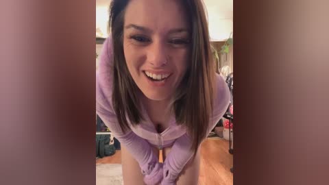 Media: Video of a smiling young woman with straight brown hair, light skin, and a nose piercing, wearing a purple hoodie, bending forward in a cozy, cluttered room with wooden floors and furniture.