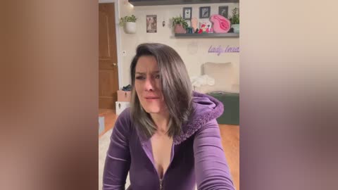 Media: A video of a young woman with shoulder-length dark hair, wearing a purple hoodie, standing in a cozy bedroom with floral decor, potted plants, and a bed.