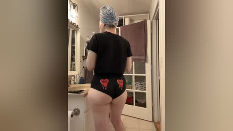 Media: Video of a woman with pale skin, wearing a black crop top and black thong with red heart designs. She has a blue headscarf and stands in a narrow hallway with beige walls and a white door.