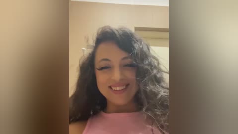 Media: Video of a smiling woman with curly black hair, wearing a pink top, standing in a narrow hallway with beige walls and a wooden door.