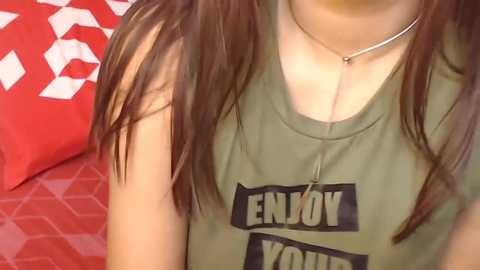 Media: Video of a young woman with long, straight brown hair, wearing a green tank top with \"ENJOY YOURSELF\" in bold black letters. She has a silver necklace and sits on a red couch with a geometric pattern.