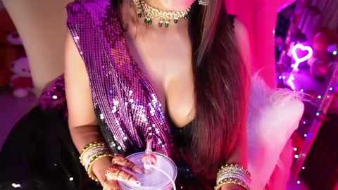 Media: Video of a woman with long, straight hair, wearing a glittery, purple sequined sari, gold jewelry, and a matching choker, holding a glass of pink liquid. The background is vibrant with neon lights and a heart-shaped cutout.