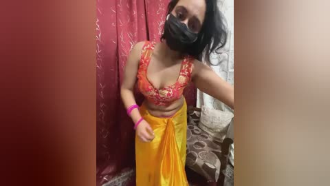 Media: Video of a South Asian woman with medium skin tone, black hair, wearing a red lace bra, yellow skirt, and black face mask, indoors with red curtains and bed visible.