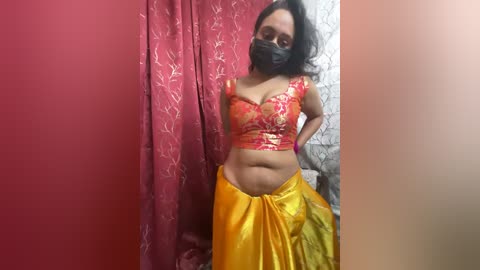 Media: A video of a middle-aged woman with a medium complexion, wearing a red and yellow saree, black face mask, and glasses, standing against a red curtain with floral patterns.