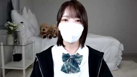 Media: Video of an East Asian woman with short, dark hair, wearing a white face mask, plaid bow tie, and black jacket, standing in a softly lit bedroom with plush toys and white bedding.