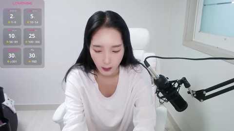 Media: Video of an Asian woman with straight black hair, wearing a white shirt, sitting at a microphone stand in a white room with a calendar and a window.