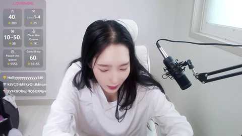 Media: Video of a young Asian woman with long black hair, fair skin, and closed eyes, wearing a white shirt, sitting at a desk with a microphone, surrounded by a white room with a digital screen displaying weather information.