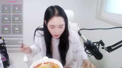 Media: Video of an East Asian woman with long black hair, wearing a white lab coat, recording a video on a white table, with a microphone and a digital clock displaying 2:55.