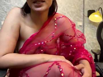 Media: A video of a South Asian woman with medium skin tone, wearing a sheer red sari adorned with sequins, sitting on a chair in a dimly lit room with grey wallpaper and a yellow lamp.