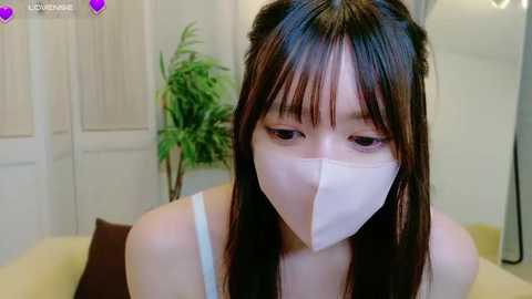 Media: A video of a young Asian woman with long black hair, wearing a white face mask, a white tank top, and sitting on a bed with a brown pillow. The background includes a green potted plant and a white door.