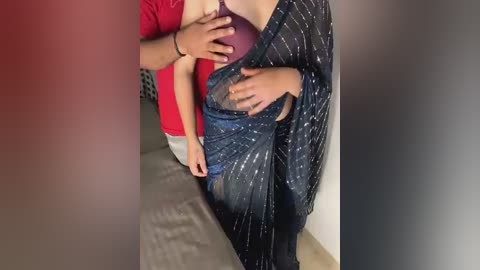 Media: Video of a woman in a black sari with white dots, holding a baby. A man in a red shirt and blue jeans stands behind her, one hand on her stomach.