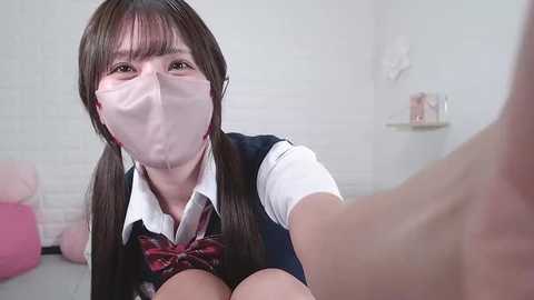 Media: Video of a young Asian woman with straight brown hair in pigtails, wearing a white face mask, navy-blue vest, and plaid tie. Background shows a white brick wall and a pink cushion.