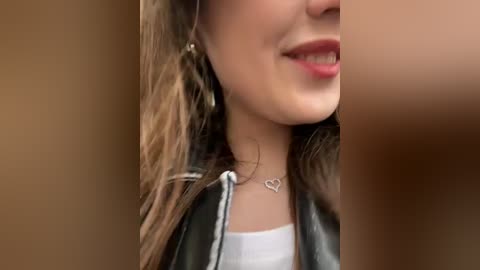 Media: A close-up video of a smiling woman with light skin, wearing a black leather jacket over a white shirt, adorned with a delicate heart pendant necklace.