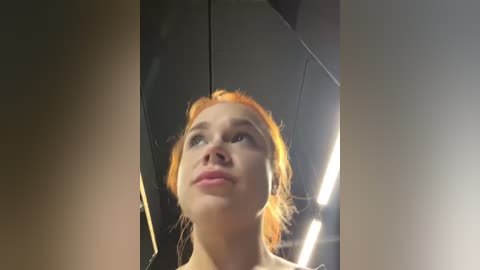 Media: Video of a young Caucasian woman with fair skin and red hair in a messy bun, gazing upward. She wears a light pink top. Background includes a dimly lit, modern interior with metallic panels and fluorescent lighting.