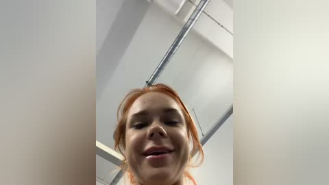 Media: Video of a young woman with fair skin and reddish-brown hair, smiling broadly, taken from a low angle. She is indoors, possibly in a bathroom or small room, with white walls and metallic fixtures in the background.