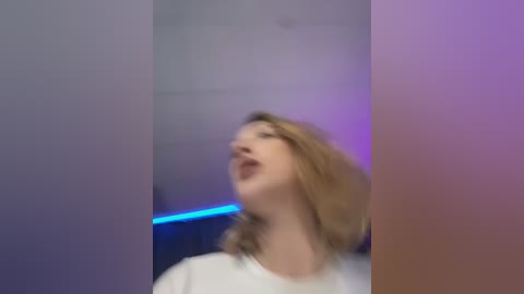 Media: Video of a fair-skinned, blonde woman with shoulder-length hair, wearing a white top, singing with eyes closed and mouth open, in a dimly lit room with blue and purple lighting.