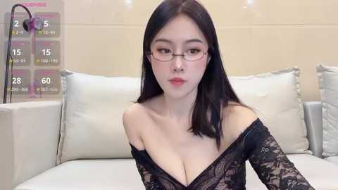 Media: A video of an East Asian woman with straight black hair and glasses, wearing a black lace off-shoulder top, sitting on a white couch. The background includes a beige wall and a calendar.