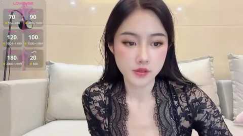 Media: Video of an East Asian woman with long black hair, fair skin, and a delicate facial structure, wearing a black lace top, sitting on a white sofa.