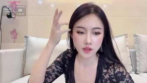 Media: Video of an East Asian woman with long black hair, wearing a black lace robe, giving a thumbs-up gesture. She is indoors, in a modern bedroom with white furniture and a beige wall.