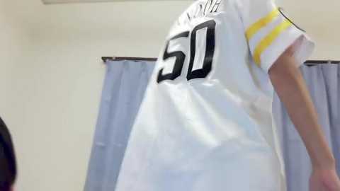 Media: Video of a white person wearing a white football jersey with black numbers 50, yellow stripes, and \"WISCONSIN\" on the back, standing in a room with light blue curtains and beige walls.