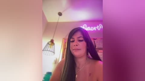 Media: Video of a topless woman with long dark hair, wearing a silver necklace, in a softly lit room with a pinkish hue, neon sign, and hanging lamp.