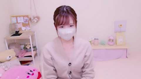 Media: A video of a young Asian woman with straight brown hair, wearing a beige cardigan, white face mask, and sitting in a pastel-colored room with a white desk and soft toys.