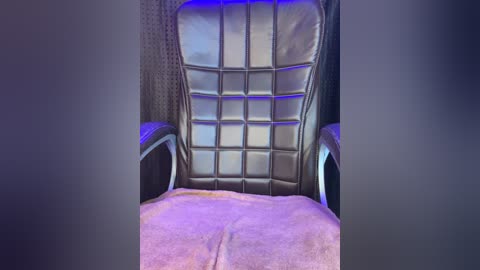 Media: A video of a modern gaming chair with a padded backrest and armrests, covered in black leatherette. The chair sits on a soft, pinkish-purple carpet. The background is dark and slightly blurred, enhancing the focus on the chair.