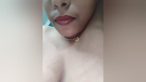 Media: A close-up video of a woman's upper torso, focusing on her chest and lips. She has light skin, full lips painted in pink lipstick, and a gold necklace with a pendant.