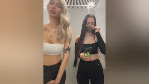 Media: Video of two young women in a modern bathroom. The blonde, wearing a white crop top and high-waisted jeans, has tattoos. The brunette, in a black crop top and leggings, holds a green drink, wearing a black mask.