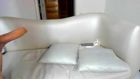 Media: Video of a white leather sectional sofa with a hand holding a smartphone. Two white cushions are placed on the sofa. Background shows a wooden dresser and open doorway.