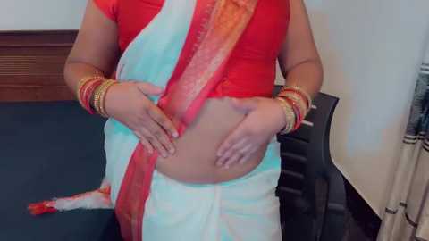 Media: Video of a woman wearing a vibrant red blouse and a white sari with red and orange borders, adorned with gold bangles. She stands in a room with a dark carpet and a wooden chair.