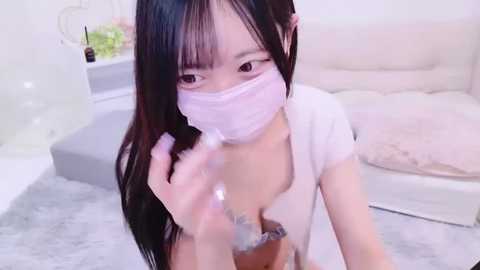 Media: A video of an East Asian woman with long black hair and pale skin, wearing a white face mask, bra, and a white shirt, kneeling on a light grey carpet in a modern, white room.