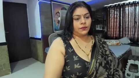 Media: Video of a South Asian woman with medium skin tone, wearing a black floral saree, seated in a modern living room with a dark wood bed, beige curtains, and a TV.