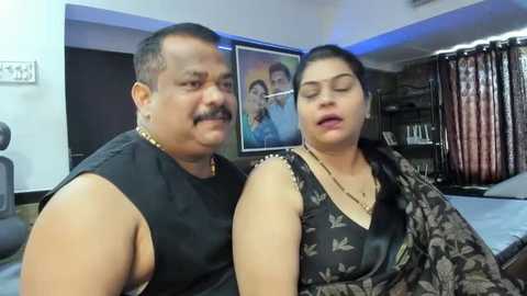 Media: Video of an Indian couple, a man with a mustache in a black sleeveless shirt, and a woman in a black floral blouse, both smiling, in a modern living room with dark curtains and a framed family photo.