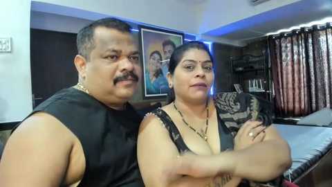 Media: Video of an Indian couple in a cozy living room; man with mustache and gold chain, woman with dark hair, both dressed casually.