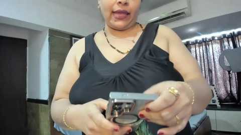 Media: Video of a middle-aged woman with light skin, wearing a black sleeveless top, gold necklaces, and a gold ring, holding a silver camera in a modern living room with air conditioning and dark curtains.