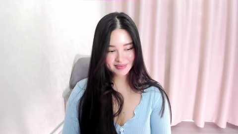 Media: A video of a smiling East Asian woman with long, straight black hair, wearing a light blue, textured cardigan, seated against a white wall and pink curtain background.