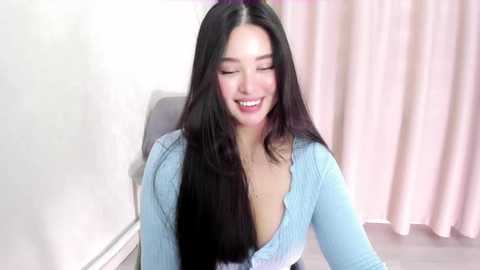 Media: A video of a smiling, young Asian woman with long black hair, wearing a light blue, ribbed, V-neck top, sitting indoors with a white wall and pink curtain in the background.