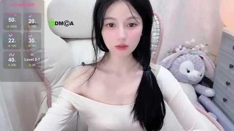 Media: Video of a young East Asian woman with long black hair, pale skin, and light makeup, wearing a white off-shoulder top, sitting on a white chair. Background includes a plush toy and a white dresser.