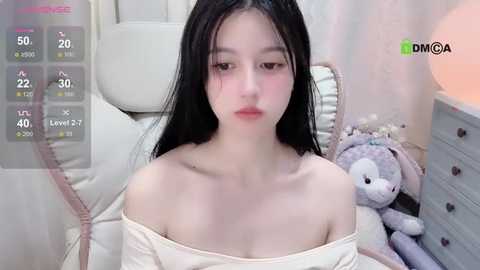 Media: A video of an East Asian woman with long black hair, wearing a beige off-shoulder top, sitting on a bed with a teddy bear beside her. The room has a soft, pastel color palette.